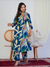 Woman wearing a blue floral dress standing in a room with a white wall and wooden chair.