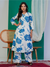 Woman wearing a white outfit with blue floral patterns in a room with a green wall and wooden chair.