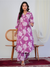 Woman wearing a purple floral outfit standing in a room with a chair and curtain.