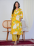 Woman wearing a yellow floral outfit standing in a room with a white curtain and wooden chair.